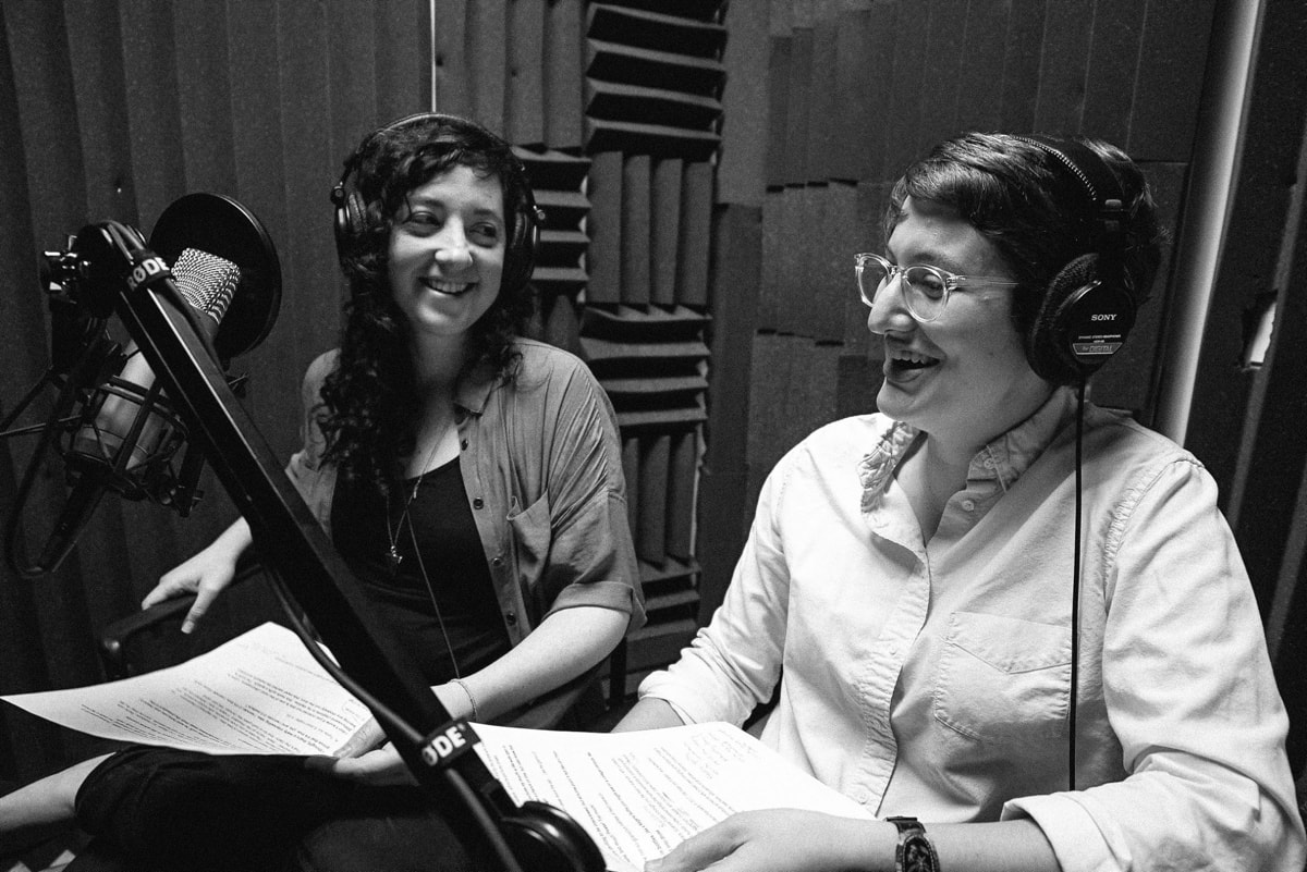 Elah and Annie with headphones and a mic in a studio with soundproofed walls.