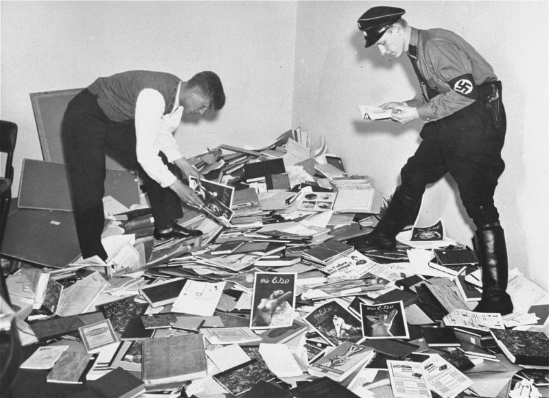 A German student and Nazi officer sift through a gigantic pile of books and photographs.