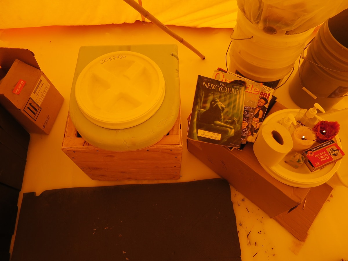 Inside a tent, a makeshift wooden toilet next to New Yorker magazines and paper plate of candles and toilet paper on top of a cardboard box.